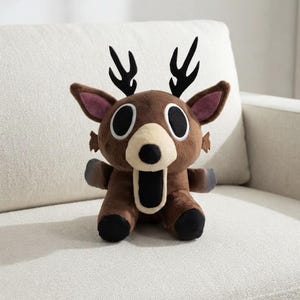May include: A brown plush deer toy featuring large black oval eyes, a cream-coloured muzzle, and a wide open black mouth. The stuffed animal has black antlers, pointed ears with purple inner lining, and dark grey accents on the paws and feet.