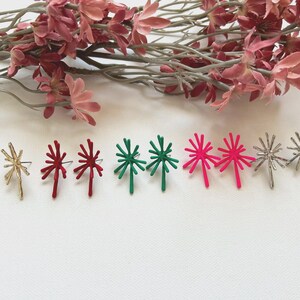 Puede incluir: Una colección de pendientes de estrella en oro, rojo, verde, rosa y plata. Los pendientes están dispuestos en fila sobre una superficie blanca, con flores rosas en el fondo. Un accesorio elegante.
