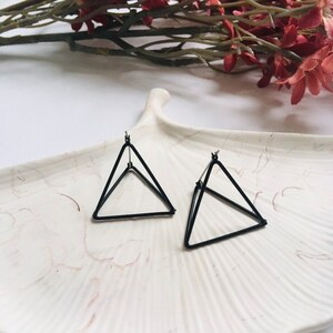 May include: A pair of black earrings featuring a three-dimensional triangular wire frame design. Each earring is constructed from thin black metal wire formed into a geometric pyramid shape, with a post backing attached at the top vertex.
