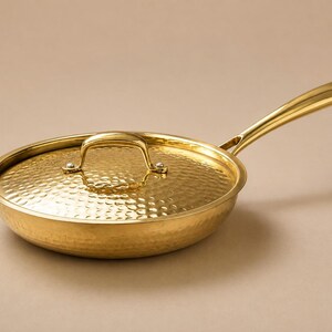 Puede incluir: Una sartén de metal en tono dorado pulido con tapa a juego, ambas con una textura martillada distintiva. La sartén incluye un mango largo y delgado y un asa curva en la tapa, mostrando un acabado metálico reflectante en todo el diseño.