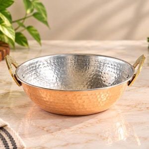 May include: A round, hammered copper serving bowl with a silver-toned interior and two decorative gold-colored metal handles attached to the sides. The bowl features a textured, dimpled surface on the exterior and a smooth, reflective metallic finish.