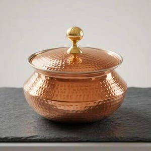 May include: A round, hammered copper serving bowl with a matching lid. The bowl features a wide, curved body and a polished, metallic finish. A small, spherical brass-colored knob sits atop the center of the lid, providing a contrast in color.