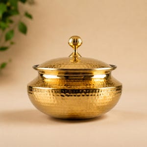 May include: A polished gold-colored metal serving bowl with a matching lid. The bowl features a distinct hammered texture across its rounded body and a small, spherical knob handle on top of the lid, creating a reflective and metallic appearance.
