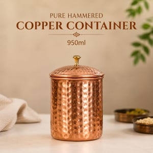 Könnte beinhalten: Ein zylindrischer Behälter aus gehämmertem Kupfer mit metallischem Glanz und einem passenden Deckel, der mit einem kleinen goldfarbenen Knauf versehen ist. Der Text PURE HAMMERED COPPER CONTAINER 950ml steht in brauner Serifenschrift darüber.