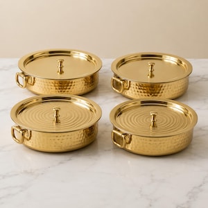 May include: Four round gold-colored metal serving dishes with lids. Each dish features a hammered texture on the sides, two small side handles, and a central knob on the lid. The lids have a concentric circular pattern radiating from the center.
