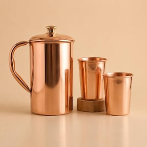 Puede incluir: Una jarra de cobre pulido con tapa y asa curva, acompañada de dos vasos de cobre a juego. La jarra presenta un cuerpo cilíndrico y un pequeño pomo en la tapa, mientras que los vasos tienen un acabado metálico liso y reflectante.