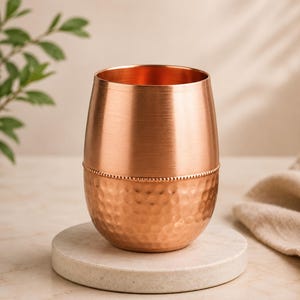 Könnte beinhalten: Ein Kupferbecher mit einem glatten oberen Teil und einem gehämmerten unteren Teil, die durch ein feines Perlenband getrennt sind. Das Gefäß hat eine abgerundete, fassartige Form mit einer metallischen Oberfläche, die das Licht reflektiert.