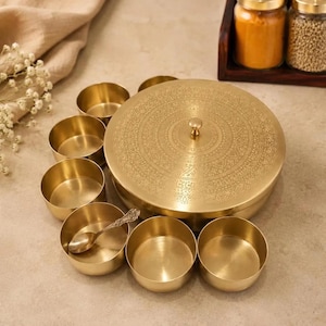 Könnte beinhalten: Eine runde Gewürzdose aus Messing mit einem aufwendig gravierten Deckel und einem zentralen Knopf, begleitet von sieben kleinen passenden Messingschalen und einem kleinen Messinglöffel. Das Set hat ein poliertes Metallfinish und ein kreisförmiges Muster.
