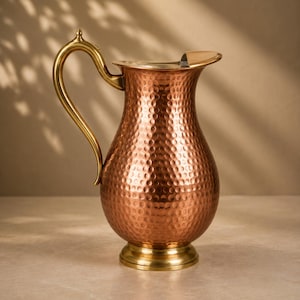 May include: A copper-coloured pitcher with a hammered texture, featuring a curved handle and a flared spout. The vessel has a rounded body that tapers towards the neck and sits on a circular, smooth, brass-toned base that matches the handle.
