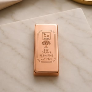 May include: A rectangular copper bar with a polished finish. The front features an engraved periodic table symbol for copper, a tree icon, and the text 500 GRAMS 99.9% FINE COPPER. The bar has rounded edges and a smooth, reflective metallic surface.