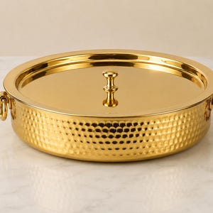 Könnte beinhalten: Eine runde, goldfarbene Servierschale mit passendem Deckel und einem zentralen Knaufgriff. Die Außenseite der Schale weist eine gehämmerte Textur auf, während die Seiten mit zwei kleinen, kreisförmigen Ringgriffen an gegenüberliegenden Seiten versehen sind.