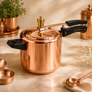 Könnte beinhalten: Ein polierter Schnellkochtopf aus Kupfer mit glänzendem Metallic-Finish, ausgestattet mit einem goldfarbenen Druckreglerventil auf dem Deckel und zwei schwarzen, hitzebeständigen Griffen. Der Seitengriff trägt die Gravur Indian Copper House.