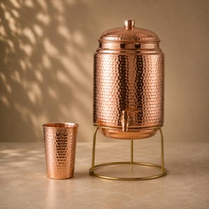 May include: A hammered copper water dispenser with a matching lid and a metal spigot, positioned on a gold-toned wire stand, accompanied by a single hammered copper tumbler. The set features a consistent metallic finish and textured surface.