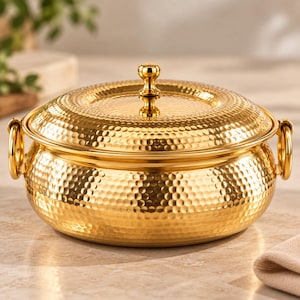 May include: A round, gold-colored serving dish with a hammered texture finish. The dish features a matching lid with a central knob handle and two circular side handles attached to the main body. The surface has a reflective, metallic sheen.