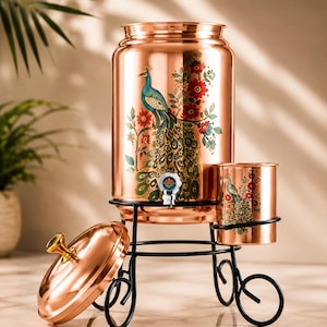 Puede incluir: Un dispensador de agua de cobre y un vaso a juego sobre un soporte de metal negro. El dispensador cilíndrico presenta un estampado colorido de un pavo real y flores con un grifo plateado. Una tapa de cobre con un pomo dorado se sitúa al lado.