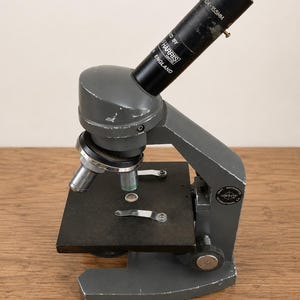 Puede incluir: Un microscopio monocular de metal gris con un ocular negro etiquetado como W10X-155MM y Harris England. El dispositivo cuenta con una platina cuadrada negra con dos clips metálicos, una torreta de objetivos giratoria y un logo de Swift.
