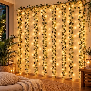 May include: A decorative curtain of artificial green ivy vines intertwined with warm white LED string lights. The vertical strands hang from a top horizontal bar, creating a glowing backdrop with realistic leaf patterns and soft illumination.
