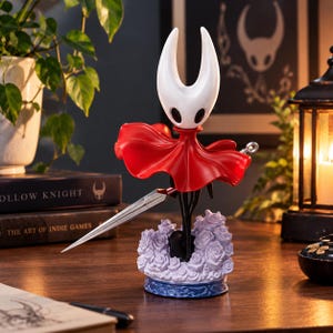 Może przedstawiać: Kolekcjonerska figurka postaci Hornet z gry Hollow Knight, z białą głową z rogami, powiewającą czerwoną peleryną i srebrną bronią w kształcie igły. Figurka stoi na podstawie z wyrzeźbionych jasnofioletowych róż z niebieskim obrzeżem.