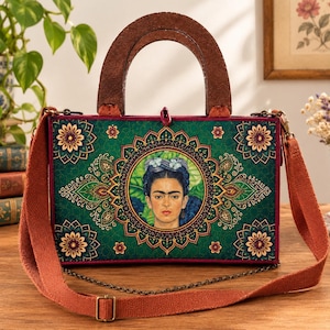 May include: A rectangular handbag featuring a portrait of Frida Kahlo centered within a green mandala pattern. The bag has a dark brown wooden handle, a detachable orange fabric shoulder strap, and a thin bronze-toned metal chain accent.
