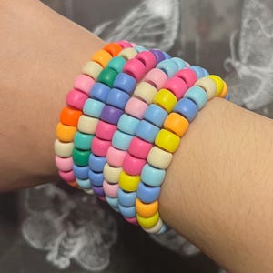 May include: A multi-strand bracelet composed of numerous small, cylindrical plastic beads in a vibrant array of colours including orange, pink, yellow, light blue, purple, green, and cream, arranged in a stacked, circular formation around the wrist.