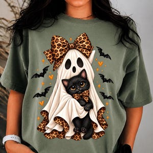 Peut inclure: Un t-shirt vert olive avec un motif représentant un chaton noir portant un nœud papillon à motif léopard, enveloppé dans un drap de fantôme blanc avec un grand nœud léopard. Le design inclut des petits cœurs orange, des étoiles et des chauves-souris.