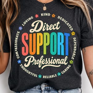 Peut inclure: Un t-shirt gris anthracite foncé arborant un motif circulaire avec le texte Direct SUPPORT Professional en grandes lettres colorées. Le texte environnant inclut les mots Loving, Kind, Dedicated, Dependable, Caring, Reliable, Thoughtful et Compassionate, agrémenté de petites icônes de fleurs colorées.