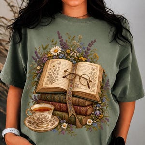 Peut inclure: Un t-shirt vert sauge avec une impression graphique représentant un livre ouvert posé sur une pile de deux livres fermés. Une paire de lunettes rondes à monture métallique repose sur les pages. Une tasse à thé fleurie est placée en dessous.