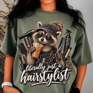May include: An olive green t-shirt featuring a graphic of a raccoon wearing a baseball cap and sunglasses, holding an iced coffee. The raccoon is surrounded by hairstyling tools including scissors, a hair dryer, and brushes. Text reads: literally just a hairstylist.