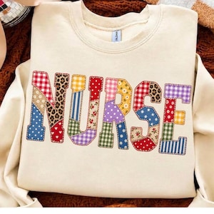 Peut inclure: Un sweat-shirt à col rond de couleur crème affichant le mot NURSE en grandes lettres style bloc. Chaque lettre est remplie d'un patchwork de divers motifs de tissu, incluant du vichy, des pois, des imprimés floraux et des taches de léopard.
