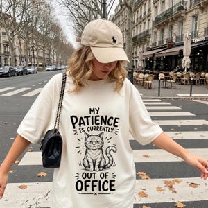 Peut inclure: Une casquette de baseball beige avec un petit logo sombre et un t-shirt crème oversize arborant un dessin noir de chat grincheux avec le texte "MY PATIENCE IS CURRENTLY OUT OF OFFICE" et un sac à bandoulière noir matelassé à chaîne.
