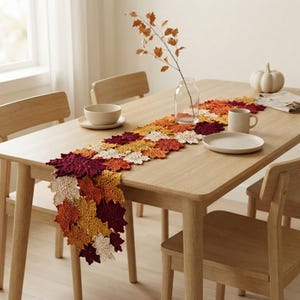 May include: A crochet table runner composed of interconnected maple leaf motifs in shades of deep burgundy, burnt orange, golden yellow, and cream. The runner is draped across a light wood dining table, accompanied by a glass vase with a branch.