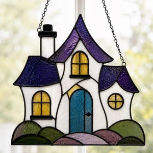 May include: A stained glass suncatcher shaped like a house with a white facade, a blue door, and purple roofs. The house features yellow windows, a black chimney, and a base of green and purple glass hills, all suspended by a black metal chain.