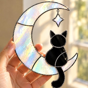 Puede incluir: Un atrapasoles de vidrieras con una luna creciente hecha de vidrio iridiscente y nacarado, y un gato de vidrio negro sólido sentado sobre la luna. Una pequeña estrella de cuatro puntas hecha de vidrio iridiscente cuelga de la luna creciente.