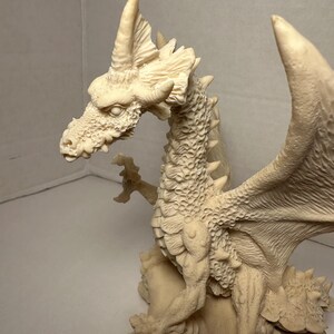May include: A cream-colored dragon figurine featuring intricate scale textures across its body, a prominent horn on its head, and large, detailed wings. The creature is depicted in a standing position with its claws extended, resting upon a base.