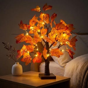 May include: A decorative tabletop tree lamp featuring artificial orange and yellow maple leaves illuminated by small warm white LED lights. The tree has a dark brown trunk and a square base, standing next to a small white ceramic vase.