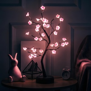 Puede incluir: Una lámpara decorativa de árbol LED con ramas de color marrón oscuro adornadas con luces brillantes en forma de flor de cerezo rosa. La lámpara tiene una base circular negra y emite una iluminación suave y cálida, creando un contraste marcado.
