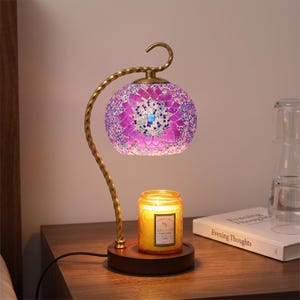 Può includere: Una lampada scaldacandela con un braccio in metallo color ottone curvo e ritorto e una base rotonda in legno. Il paralume è un globo sferico in vetro a mosaico con motivi geometrici viola, rosa e blu. Sotto è posizionata una candela gialla.