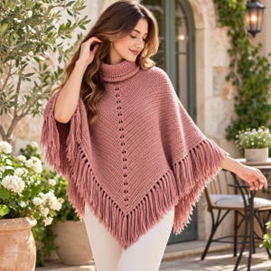May include: A dusty rose crochet poncho featuring a high turtleneck collar, a V-shaped textured knit pattern down the center, and long fringe trim along the bottom edges. The garment is constructed with a thick yarn and has a pointed hemline.