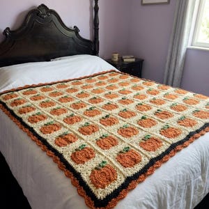 May include: A rectangular crochet blanket featuring a repeating grid pattern of orange pumpkins with green stems on a cream-colored background. The blanket is finished with a decorative black and orange scalloped border along the outer edge.