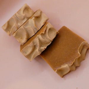 Peut inclure: Trois barres de savon rectangulaires de couleur beige disposées en pile diagonale. Chaque barre présente une surface lisse et solide avec un bord texturé et tourbillonnant sur un côté, affichant une teinte brun clair uniforme sur l'ensemble.