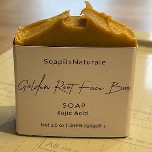May include: A rectangular bar of golden-yellow soap with a textured top edge, wrapped in a white paper label. The label reads SoapRxNaturale, Golden Root Face Bar, SOAP, Kojic Acid, and Net 4.6 oz | GRFB 290926-1, positioned on a cutting mat.