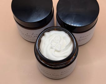 Può includere: Tre barattoli in vetro ambrato con coperchi neri contenenti burro corpo montato. Il barattolo centrale è aperto e rivela una crema bianca a spirale. Ogni barattolo presenta un'etichetta bianca con il testo Whipped Body Butter.
