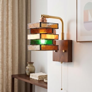 Può includere: Una lampada da parete con base rettangolare in legno e braccio in ottone che sostiene un paralume in vetro colorato a più strati. Il paralume è composto da pannelli di vetro sovrapposti nei toni dell'ambra, del bianco e del verde.