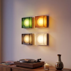 Op de afbeelding: Vier vierkante wandlampen met glazen kappen in groen, amber, donkergrijs en transparant, die elk een warme, cirkelvormige gloed uitstralen. De lampen zijn in een twee-bij-twee raster op de muur geplaatst en geven zacht licht aan de kamer.