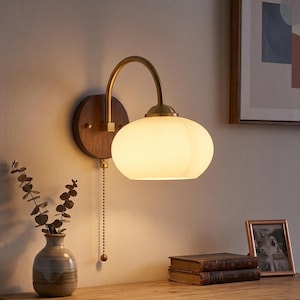May include: A wall-mounted lamp featuring a circular dark wood base, a curved brass arm, and a rounded, frosted white glass shade. The fixture includes a brass pull chain switch hanging vertically from the base, emitting a warm, soft glow.