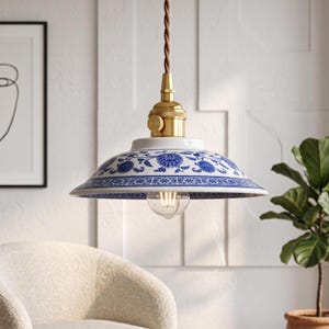 Op de afbeelding: Een hanglamp met een brede, ondiepe keramische kap versierd met een blauw-wit bloemmotief. Het armatuur is voorzien van een goudkleurige metalen fitting en een gedraaid bruin stoffen snoer, met een zichtbare transparante gloeilamp eronder.