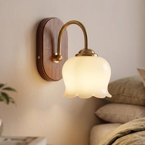 Può includere: Una lampada da parete con una piastra posteriore ovale in legno marrone scuro, un braccio curvo in ottone e un paralume in vetro bianco smerigliato a forma di campana. L'apparecchio emette una luce soffusa e il bordo del vetro ricorda un petalo.