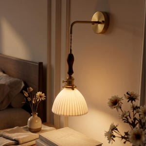 May include: A wall-mounted lamp featuring a circular brass base, a curved brass arm, and a dark brown wooden handle. The light fixture includes a white, ribbed, bell-shaped glass shade that emits a warm, soft glow, suspended by a twisted cord.
