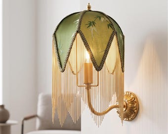 May include: A wall-mounted lamp featuring a green fabric shade with a bamboo leaf pattern and a scalloped edge. The shade is adorned with long, dangling gold-toned metal chains. The lamp has a curved gold-toned metal arm and a circular wall mount.