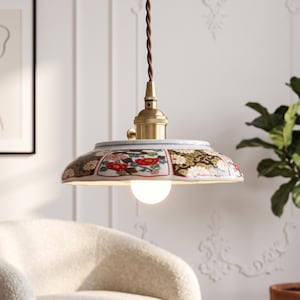 May include: A pendant light featuring a wide, shallow ceramic shade decorated with a colourful floral pattern in red, white, and green. The fixture includes a brass-toned metal socket and a brown twisted fabric cord, suspending a single light bulb.